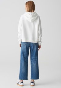 Felpa bianca con una vestibilità rilassata, polsini a costine e cappuccio con lacci, abbinata a jeans blu a gamba larga e sneaker bianche con accenti blu navy.