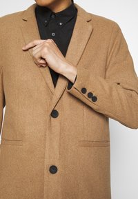 Cappotto di lana beige con revers a notched, design monopettino, tre bottoni neri e tasche laterali, indossato sopra una camicia nera.