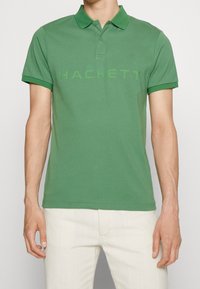 Polo en coton vert avec un col classique, des manches courtes et le mot "HACKETT" imprimé en vert clair sur le devant.