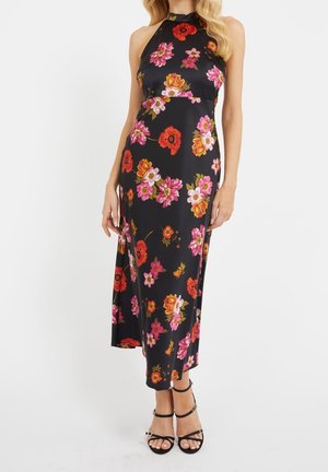 Femme portant une robe noire sans manches à col halter avec un imprimé floral rose, rouge et orange, associée à des sandales noires à talons hauts avec des lanières.