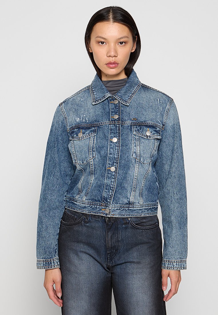 Chaqueta de mezclilla recortada en azul claro con cuello clásico, cierre frontal de botones y dos bolsillos en el pecho. Tejido texturizado con acentos desvaídos.