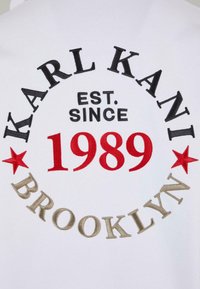 Felpa bianca con testo ricamato circolare: "KARL KANI," "EST. SINCE," "1989," e "BROOKLYN," con accenti rossi e dorati.