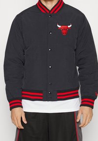 Schwarze Bomberjacke mit rot gestreiften Bündchen und Saum, ausgestattet mit einem roten, gestickten Bullen-Logo auf der Brust und Druckknopfverschluss.