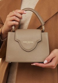 Des mains tenant un petit sac à main taupe texturé avec une poignée supérieure et un logo doré "Isabel Bernard Paris" sur le rabat avant.