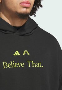 Sort hoodie med lysende gul tekst, der står "Tro på det." Inkluderer to logo-designs over teksten. Blødt, tekstureret stof.