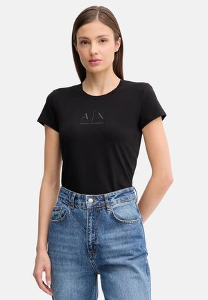 Donna che indossa una maglietta nera aderente Armani Exchange e jeans blu a vita alta, in piedi con una mano dietro la schiena.