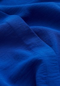 Kungblått tyg med en slät textur, präglat av subtila veck och synliga sömmar som framhäver dess mjukhet och fall.