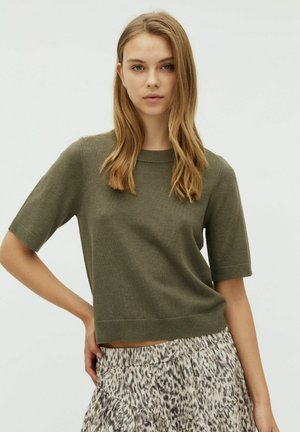 Junge Frau mit glatten blonden Haaren, die ein kurzärmeliges, olivgrünes Strickoberteil und einen gemusterten, fließenden Rock trägt, die Hand auf der Hüfte ruhend.