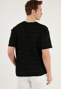 Juoda megzta T-shirt su vertikaliu raukšlėtu tekstūru, trumpomis rankovėmis ir apvaliu iškirpimu, pasižyminti laisvu siluetu ir subtiliomis permatomomis vietomis.