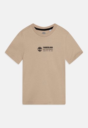 T-shirt en coton beige avec un col rond, des manches courtes et un logo Timberland noir imprimé sur le haut à gauche de la poitrine.