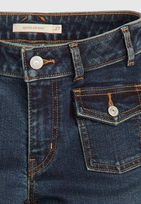 Jean en denim bleu foncé avec surpiqûres orange, bouton métallique à l'avant, passants de ceinture et une petite poche à rabat avec un bouton. Étiquette de taille 27.