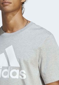 Šedé bavlněné tričko s velkým bílým logem Adidas na přední straně. Tričko má kulatý výstřih a krátké rukávy.