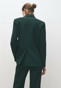 Ciemnozielona marynarka o dopasowanym kroju, z klapą typu szczytowego i rozcięciem z tyłu, wykonana z teksturowanej tkaniny. Zrównoważona, smukła sylwetka.