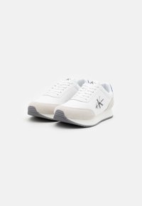 Zapatos deportivos blancos con una parte superior de tela suave y acentos de ante gris claro en la puntera. Presentan cordones, suelas de goma y detalles del logo.