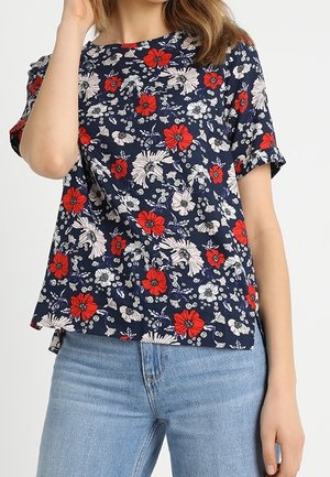 Vrouw draagt een marineblauwe blouse met korte mouwen met rood-witte bloemmotieven, gecombineerd met lichtblauwe spijkerbroek.