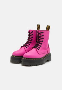 Stivali da combattimento in pelle rosa brillante con design di lacci neri, suola nera spessa e cuciture gialle lungo il bordo. Presenta un inserto per il tallone.