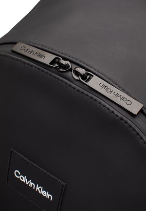 Zaino nero realizzato in materiale morbido, con una forma curva, una patch con logo in rilievo e hardware della cerniera metallica con dettaglio del marchio inciso.