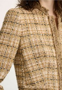 Texturovaná zlatá a béžová bunda s mřížkovým vzorem a černobílými akcenty. Tkanina má mírně třpytivý povrch a vysoký límec.