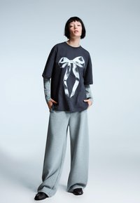 Sort t-shirt med en hvid sløjfegrafik, parret med løse grå sweatpants og mørke sko. Modellen har kort hår og hænderne i lommerne.