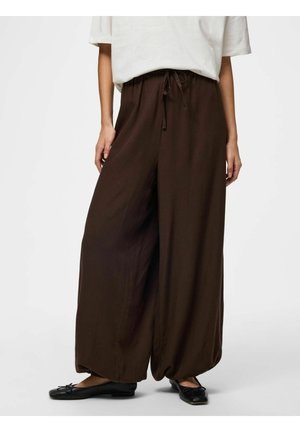 Osoba ubrana w luźne, ciemnobrązowe, szerokie spodnie z sznurkiem w pasie, połączone z czarnymi balerinami i białą bluzką.