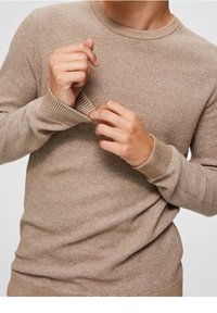 Pull en tricot beige avec un motif texturé, encolure ronde et poignets côtelés. Le tissu semble doux et léger avec une coupe décontractée.