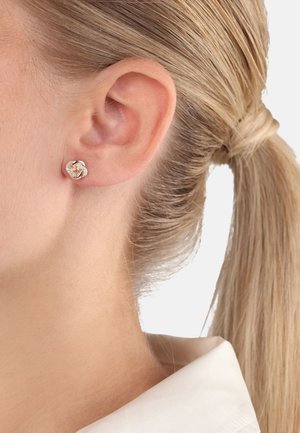 Pendientes de tuerca en oro rosa con un diseño retorcido y acentos transparentes, mostrados en la oreja de un modelo con cabello rubio claro.