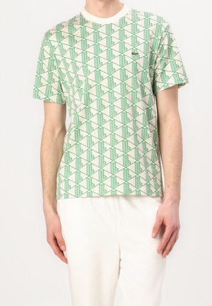 Homme portant un t-shirt blanc à col rond avec un motif géométrique vert et un petit logo crocodile sur la poitrine, associé à un pantalon blanc.