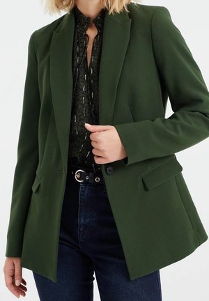 Kvinde iført mørkegrøn blazer over sort mønstret bluse og mørke jeans, holder blazerlap med den ene hånd.