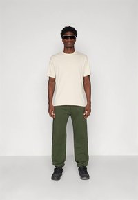 Mężczyzna ubrany w beżową koszulę z krótkim rękawem, oliwkowe spodnie, czarne sneakersy i czarne okulary przeciwsłoneczne stoi na tle białego tła.