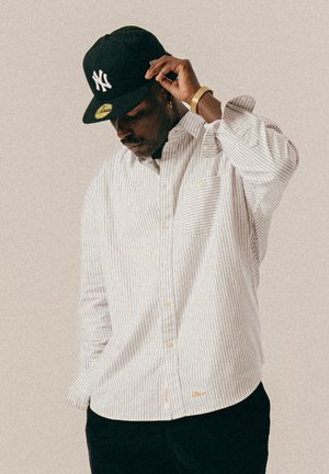 Mann mit einer schwarzen New York Yankees-Cap, goldener Uhr, Creolen, einem beigen gestreiften Hemd mit Knöpfen und schwarzen Hosen, der mit leicht gesenktem Kopf posiert.