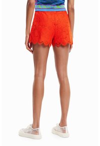 Shorts en dentelle florale orange avec un ourlet festonné, présentant une texture lisse et une coupe ajustée, associés à des baskets transparentes.
