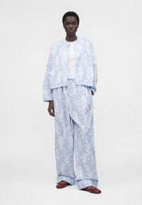 Γυναίκα που φοράει ανοιχτό μπλε ζακέτα με σχέδιο και φαρδιά παντελόνια που ταιριάζουν, λευκό τοπ και μπορντό επίπεδα παπούτσια, στέκεται σε λευκό φόντο.