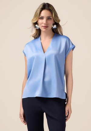 Hemelsblauwe blouse met korte mouwen, een V-hals en subtiele plooidetails. Gladde stof met een ontspannen pasvorm, gecombineerd met donkere broeken.