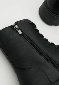 Botas de cuero negro hasta el tobillo con cremallera frontal, punta redonda y suela de goma texturizada con detalle de cordones adicional.