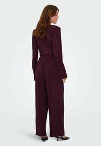 Bodysuit plissé bourgogne avec manches longues, jambes larges et taille ajustée. Tissu lisse avec une silhouette raffinée. Chaussures à talons noires.