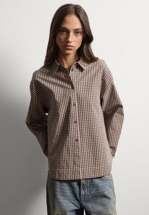 Young woman with long dark hair wears a brown and beige checkered button-up shirt and light blue jeans against a plain background.