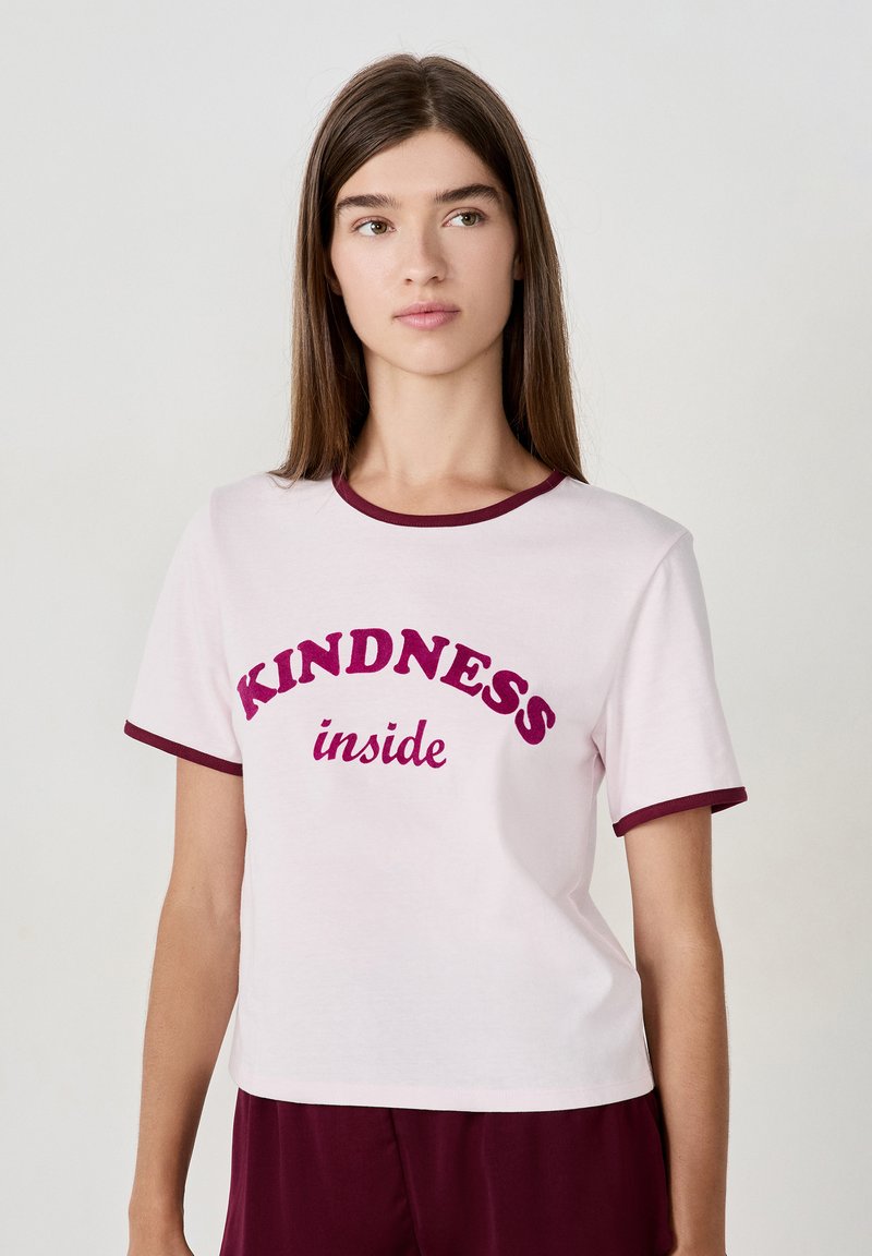 Giovane donna con lunghi capelli castani indossa una maglietta bianca con bordi bordeaux e la scritta "KINDNESS inside" in lettere grasse.