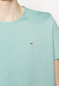 Světle modré bavlněné tričko s kulatým výstřihem. Má malou vyšívanou značku na hrudi v bílé, červené a modré barvě. Hladká textura.