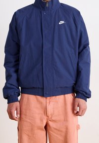 Marinblå vindjacka med hög krage, dragkedjestängning, elastiska ärmslut och Nike-logotyp på vänster bröst. Har sidofickor.