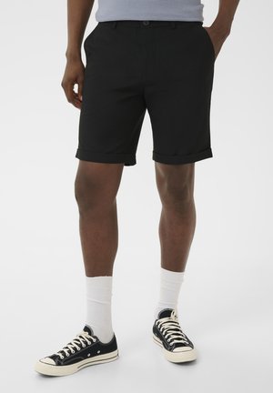 Homme portant un short noir à revers roulé, chaussettes blanches montantes, baskets basses noires, debout avec une main dans la poche sur fond uni.