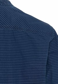 Sininen nappipaita, jossa on ohuita pystysuoria raidoituksia, kaulus ja hienovaraisia ommellut yksityiskohtia. Kangas näyttää tekstuuriltaan kevyeltä.