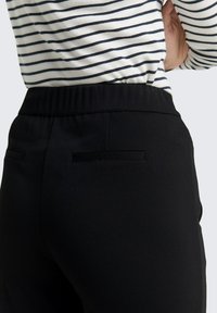 Zwarte elastische broeken met een gladde textuur. Twee kleine, elegante zakken achteraan. Draagt een wit en marineblauw gestreept bovenstuk.