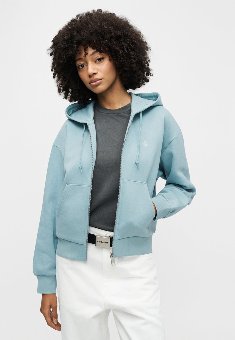 Jeune femme aux cheveux bouclés portant un sweat à capuche zippé bleu clair, un t-shirt gris foncé, un pantalon blanc et une ceinture, sur un fond blanc uni.