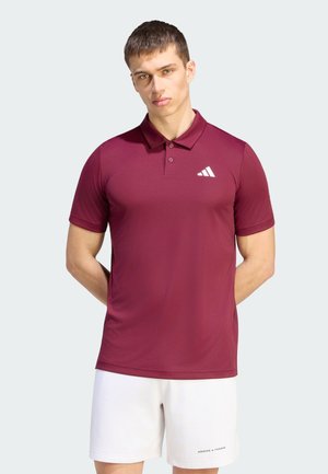 Joven con una camisa de polo color granate con un logo blanco y pantalones cortos blancos, de pie con las manos detrás de la espalda contra un fondo liso.