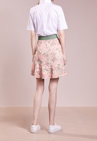 Žena v bílém krátkém rukávu tričku zasunutém do růžové květované sukně se zeleným pasem a bílými teniskami, stojící na béžové podlaze.