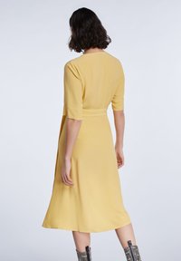 Gul klänning med figursydd midja, korta ärmar och utsvängd fåll. Slät tyg utan synliga mönster; modellen bär metalliska stövlar.
