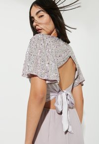Top in lavanda con maniche a cappotto decorate da paillettes e schiena scoperta, dotato di un nastro di raso. Tessuto liscio e leggero luccichio su tutta la superficie.