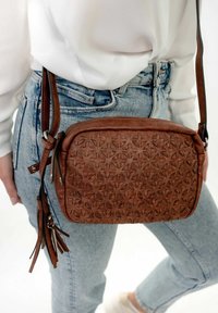 Borsa a spalla in pelle marrone con motivo floreale impresso, forma rettangolare, tracolla regolabile e superficie testurizzata, indossata sopra un tessuto in denim.