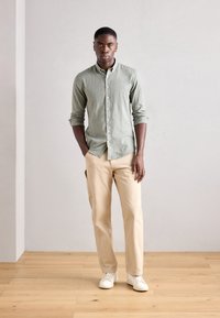 Cămașă verde deschis, cu croială relaxată, asociată cu pantaloni bej și adidași albi, stând pe un podea din lemn, într-un fundal minimalist.