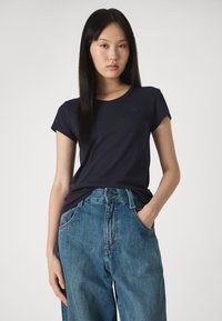 Σαλομπαλάνα μπλε χρώματος από βαμβάκι με κοντά μανίκια, στρογγυλή λαιμόκοψη και μικρό λογότυπο, συνδυασμένη με ψηλόμεσα, φαρδιά, ανοιχτόχρωμα τζιν.